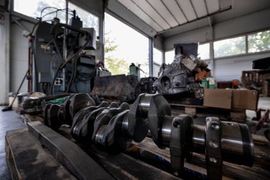 Parçalanmış bir dizel kamyon motoru işlek bir tamirhanede ya da depoda ahşap desteklerin üzerinde yatıyor ve arka planda eğimli bir kamyon taksisi görünüyor.