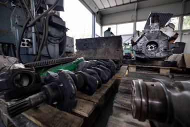 Parçalanmış bir dizel kamyon motoru işlek bir tamirhanede ya da depoda ahşap desteklerin üzerinde yatıyor ve arka planda eğimli bir kamyon taksisi görünüyor.