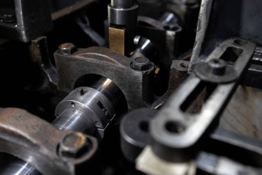 Parçalanmış bir dizel kamyon motoru işlek bir tamirhanede ya da depoda ahşap desteklerin üzerinde yatıyor ve arka planda eğimli bir kamyon taksisi görünüyor.