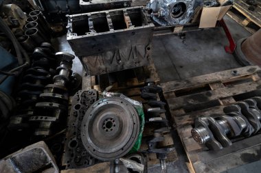 Parçalanmış bir dizel kamyon motoru işlek bir tamirhanede ya da depoda ahşap desteklerin üzerinde yatıyor ve arka planda eğimli bir kamyon taksisi görünüyor.