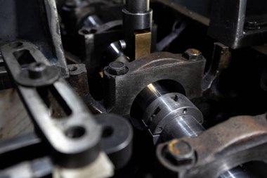 Parçalanmış bir dizel kamyon motoru işlek bir tamirhanede ya da depoda ahşap desteklerin üzerinde yatıyor ve arka planda eğimli bir kamyon taksisi görünüyor.