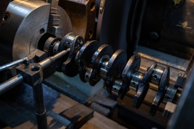 Parçalanmış bir dizel kamyon motoru işlek bir tamirhanede ya da depoda ahşap desteklerin üzerinde yatıyor ve arka planda eğimli bir kamyon taksisi görünüyor.