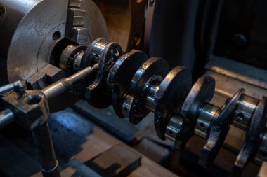 Parçalanmış bir dizel kamyon motoru işlek bir tamirhanede ya da depoda ahşap desteklerin üzerinde yatıyor ve arka planda eğimli bir kamyon taksisi görünüyor.