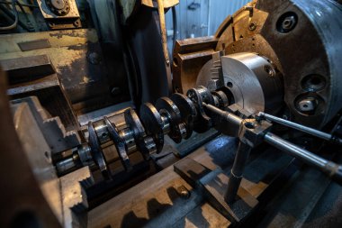 Parçalanmış bir dizel kamyon motoru işlek bir tamirhanede ya da depoda ahşap desteklerin üzerinde yatıyor ve arka planda eğimli bir kamyon taksisi görünüyor.