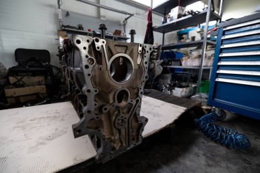 Parçalanmış bir dizel kamyon motoru işlek bir tamirhanede ya da depoda ahşap desteklerin üzerinde yatıyor ve arka planda eğimli bir kamyon taksisi görünüyor.