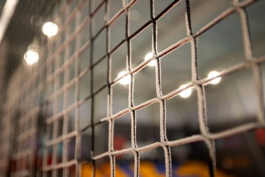 Kare şeklindeki siyah voleybol ağının kapalı bir voleybol sahasında belirsiz renkli şekilleri ve parlak ışıkları gösteren bulanık bir arka planı var..