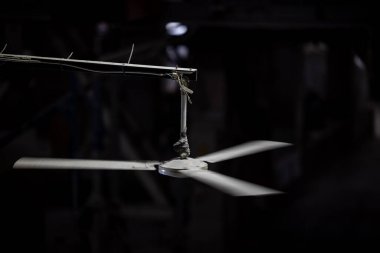 Üç pervaneli bir endüstriyel tavan pervanesi tek bir ışıkla aydınlatılan karanlık bir arka plana doğru hızla dönüyor..