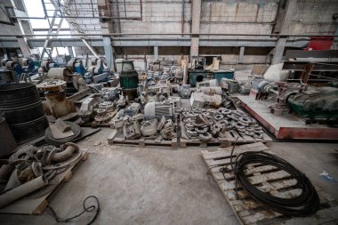 Beton zemine saçılmış elektrik motorları, pompalar, fıçılar ve makineler olan dağınık bir endüstriyel depo. Arka planda büyük mavi bir vinç göze çarpıyor..