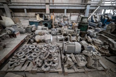 Beton zemine saçılmış elektrik motorları, pompalar, fıçılar ve makineler olan dağınık bir endüstriyel depo. Arka planda büyük mavi bir vinç göze çarpıyor..