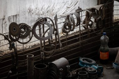 Çeşitli yağlı metal parçaları kirli bir pencereye bağlı bir tel örgüden sarkıyor ve daha fazla alet ve parça aşağıdaki tezgaha saçılmış durumda. Bir şişe sola oturur..