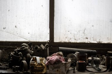 Metal parçaları olan kirli bir pencerenin önündeki dağınık tezgah: dişliler, borular, tesisatlar. Dağınık el aletleri ve eskimiş bir karton kutuda buruşmuş pembe bir bez. Meşgul, ihmal edilmiş endüstriyel atölye.