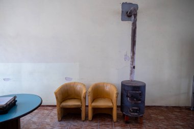 Basit bir bekleme salonunda, soluk sarı bir duvara dayanan bir baca borusuyla silindirik metal bir sobanın yanında iki aşınmış sarı sandalye yer alıyor. Mavi bir masa kısmen sağda görünür. Zemin kiremitli.