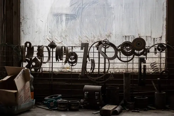 Çeşitli yağlı metal parçaları kirli bir pencereye bağlı bir tel örgüden sarkıyor ve daha fazla alet ve parça aşağıdaki tezgaha saçılmış durumda. Bir karton kutu solda duruyor..