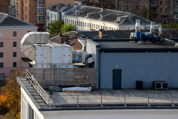 Birkaç büyük havalandırma ünitesi ve geniş bir kanal sistemi olan bir ofis binasının çatısında. Arka planda çeşitli mimari tasarımlara sahip konut ve ticari binalar yer alıyor.