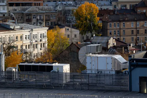 Birkaç büyük havalandırma ünitesi ve geniş bir kanal sistemi olan bir ofis binasının çatısında. Arka planda çeşitli mimari tasarımlara sahip konut ve ticari binalar yer alıyor.