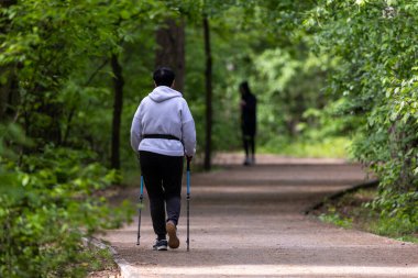 Parkta bir terrenkur patikasında yürüyen bir kadın. Spor kıyafetleri giyer ve yürüme sopaları kullanır. Çevresi yeşilliklerle çevrili olarak belirlenmiş yolda sabit bir hızda hareket eder..