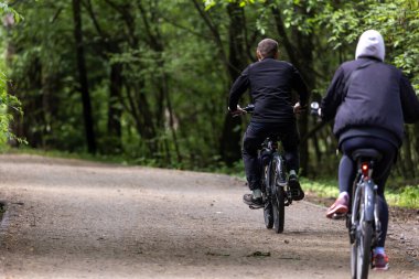Çevresi yeşilliklerle çevrili, ağaçlık bir patika boyunca giden iki bisikletçi. Siyah elbiseli kız ve erkek. Sahne, doğadaki günlük aktiviteleri ve hareketleri yansıtıyor..