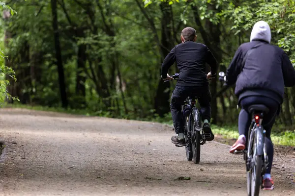 Çevresi yeşilliklerle çevrili, ağaçlık bir patika boyunca giden iki bisikletçi. Siyah elbiseli kız ve erkek. Sahne, doğadaki günlük aktiviteleri ve hareketleri yansıtıyor..