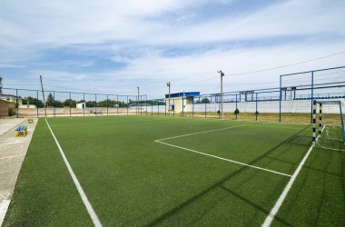 Yapay çim, beyaz sınır çizgileri ve her iki ucunda kale direkleri olan mini bir futbol sahası. Mavi ve sarı banklar yan yana, mavi metal bir çitle çevrili..