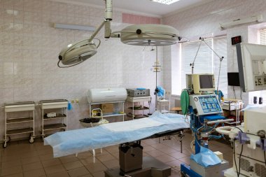 Sağ ön planda, bir video endoskopi sistemi görülebilir, merkezinde bir akciğer solunum cihazı ve cerrahi masası var. Oda temiz, çeşitli tıbbi gereçler ve cerrahi ışıklandırmayla dolu..