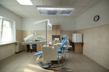 Modern bir diş sağlığı kliniğinde mavi bir dişçi koltuğu, aletleri olan beyaz bir diş ünitesi, ameliyat lambası ve temiz bir tedavi alanında ahşap bir dolap bulunur..