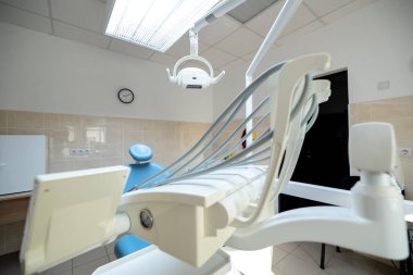 Modern bir dişçi kliniğinde mavi bir dişçi koltuğu, aletleri olan beyaz bir diş ünitesi, ameliyat lambası ve temiz bir ayarda bir duvar saati bulunur..