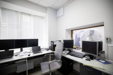 Tıbbi kontrol odasında birden fazla bilgisayar monitörü, dizüstü bilgisayar ve ofis sandalyesi var. Binanın bodrum katında bitişik bir tomografi odasına bakan bir pencere var..