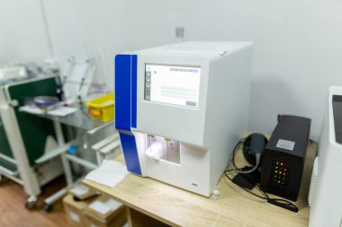 Analiz sonuçlarını gösteren dijital ekranlı bir tıbbi kan analizcisi ve klinik hastanesindeki bir klinik odasındaki ahşap bir masada duran siyah kesintisiz bir güç kaynağı..