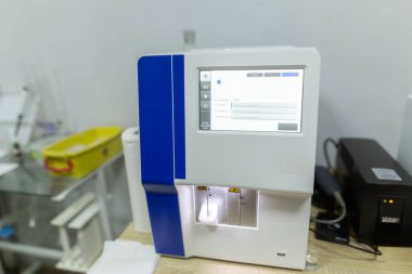 Analiz sonuçlarını gösteren dijital ekranlı bir tıbbi kan analizcisi ve klinik hastanesindeki bir klinik odasındaki ahşap bir masada duran siyah kesintisiz bir güç kaynağı..