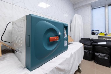 Yeni, fişi takılı olmayan bir sterilizatör, bir otoklava temiz bir hastane odasında fayanslı duvarlar ve yakınlardaki ekipman çantalarıyla dinleniyor..