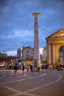 Bordeaux, Fransa - 23 Kasım 2021: Place de la Victoire 'ın akşam manzarası, merkezi bir pembe mermer sütun ve meydandaki insan ve tramvaylarla birlikte anıtsal Porte d' Aquitaine kemeri.