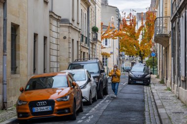 Bordeaux, Fransa - 23 Kasım 2021 Bordeaux 'da dar bir sokakta turuncu bir Audi, turuncu ceketli bir kadın, geleneksel binalar ve turuncu yapraklı bir Ginkgo ağacı da dahil olmak üzere park edilmiş arabalar yer alıyor.