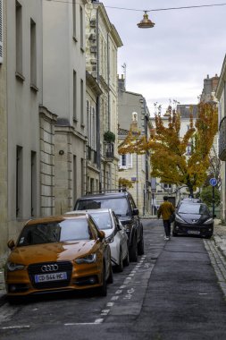 Bordeaux, Fransa - 23 Kasım 2021: Bordeaux 'da dar bir sokakta turuncu bir Audi, turuncu ceketli bir adam, geleneksel binalar ve turuncu yapraklı bir Ginkgo ağacı da dahil olmak üzere park edilmiş arabalar yer alıyor..