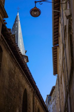 Dar bir cadde Eglise Saint-Eloi kulesine doğru gidiyor. Eski taş binalar ve mavi gökyüzünün altında antik bir sokak lambası var.