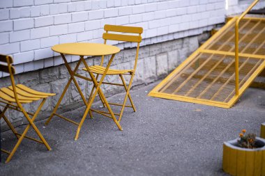 İki sarı metal sandalye ve bir yuvarlak masa asfalt üzerinde beyaz tuğla bir duvara karşı, sarı metal bir rampa ya da merdiven yanında, ön planda sarı bir varil dikici.