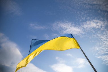 Rüzgarda dalgalanan Ukrayna bayrağı, mavi ve sarı çizgilerini gösteriyor, dağınık beyaz bulutlu mavi gökyüzüne karşı.