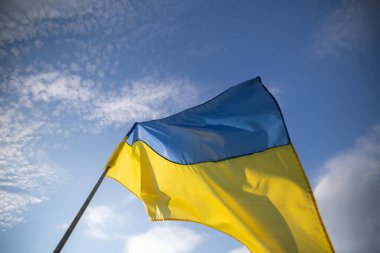 Rüzgarda dalgalanan Ukrayna bayrağı, mavi ve sarı çizgilerini gösteriyor, dağınık beyaz bulutlu mavi gökyüzüne karşı.