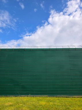 Uzun, koyu yeşil kıvrımlı metal bir duvar yeşil çimen şeridinin üzerinde yatay olarak uzanır..