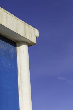 Beyaz fayanslı yüzeyleri ve mavi yansıtıcı paneli olan modern bir bina köşesi açık mavi gökyüzü ile buluşur, hafif yıpranma ve leke izlerini gösterir..