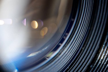 Bir kamera merceğinin soyut makrosu dokulu odak halkasını ve ön elementi gösterir. Renkli bokeh daireler ve mavi ışık parlaması çok katmanlı optik camlara yansıyor..