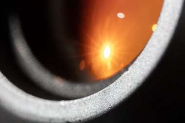 Parlak güneş patlaması yansıması olan kamera merceğinin makro görüntüsü. Turuncu kaplamalı camda, desenli metal mercek namlusuyla çevrili yıldız benzeri parlama ve bokeh görülebilir..