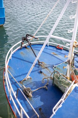 Bir balıkçı teknesinin pruvasının yıpranmış mavi güvertesi. Pas ve soyulan boyaları gösteriyor. Güvertede siyah bir çapa ve aşınmış ipler var..