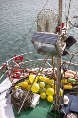 Bir balıkçı teknesinin güvertesi, sarı şamandıralar, halatlar ve ağlar da dahil olmak üzere teçhizatla doludur. Büyük bir güverte ışığı ve bir vinç görünür..