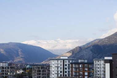 Uzakta karla kaplı dağ zirvelerinin manzarası ve ön planda yerleşim binaları. Gökyüzü açık, beyaz tepeleri vurguluyor..