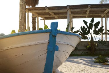 Kırsal ahşap bir tekne beyaz ve maviye boyanmış, tropikal bitkiler ve parlak güneş ışığıyla çevrili, yıpranmış ahşap bir yapının yanında duruyor..