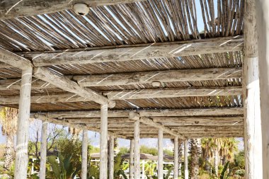 Çatıyı oluşturan kalın kirişli, taşralı ahşap bir pergola, yere gölgeler bırakıyor. Güneş ışığı kirişlerin arasından süzülerek, tahtanın doğal dokusunu vurgular..