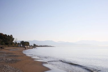 Kıyıya ulaşan yumuşak dalgalarla sakin, çakıl taşlı bir sahil. Uzak dağlar yumuşak ışıkla yıkanır, kıyı şeridi ufka doğru uzanır..