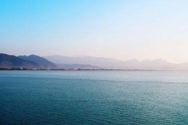 Ön planda sakin bir deniz, dağların uzak bir manzarası kısmen sisle kaplı. Açık gökyüzü açık mavi bir eğimle.