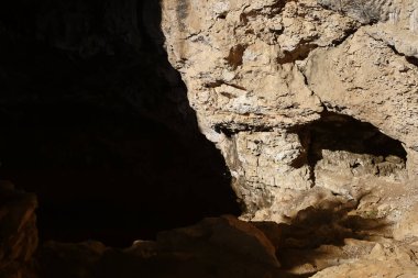 Güneş ışığıyla aydınlatılan bir mağara girişi ön planda engebeli kayalık arazi ve mağaranın iç kısımlarında gölgeler var..