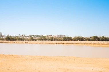 Su ve uzak binalarla çöl manzarası.